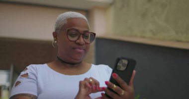 Afrika kökenli bir kadın akıllı telefon kullanıyor, iletişim kuruyor ve konsantre oluyor, gündelik dekor ve bitkilerle dolu bir konut ortamında.