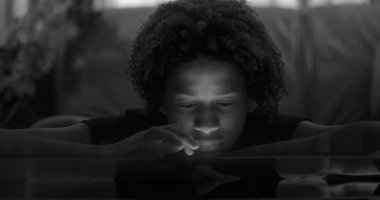 Masaya yaslanan çocuğun yakın çekimi, akıllı telefonun yüzü parlıyor, boş ifade, dijital bağımlılığı vurguluyor ve duygusal yoksunluğu, siyah beyaz