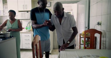 Yaşlı Afrikalı Amerikalı bir adama oğlu tarafından sıcak bir mutfakta yemek masasına oturtulması günlük hayatta bakım, şefkat ve aile bağlarını yansıtıyor.