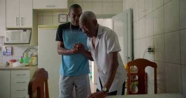 Yaşlı Afrikalı Amerikalı bir adam, bir mutfakta oğlu tarafından destekleniyor, aile bağının samimi bir anında bakım, yardım ve şefkati yansıtıyor.
