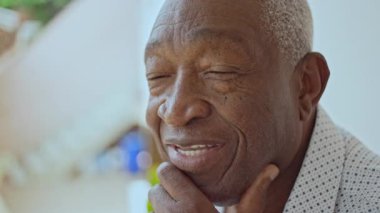 Avlunun dışında oturmuş, çenesinde bir elle gülümseyen yaşlı adamın yakın çekimi.