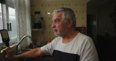 Mutfakta yaşlı bir adamın yakın çekimi, yansıtıcı bir ifadeyle dışarı bakmak, sıradan bir iç mekan anı, ev hayatı, samimi ve doğal günlük ortam.