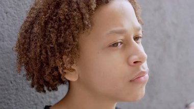 Kıvırcık saçlı, düşünceli bir ifadeyle dik dik bakan, sessiz sedasız gri duvarın yanında duran genç çocuğun yakın çekimi.