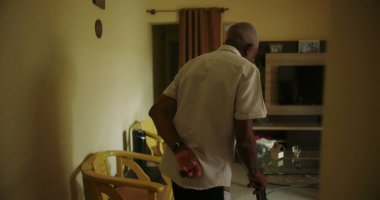 Oturma odasında elinde bastonla dolaşan yaşlı bir adam, dayanıklılık, bağımsızlık ve tanıdık bir ev ortamında yaşlanma zarafeti gösteriyor.