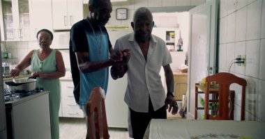 Yaşlı Afrikalı Amerikalı bir adama oğlu tarafından sıcak bir mutfakta yemek masasına oturtulması günlük hayatta bakım, şefkat ve aile bağlarını yansıtıyor.