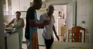 Oğlu tarafından desteklenen, koridorda yürüyen yaşlı Afrikalı Amerikalı bir adam, huzurlu bir ev ortamında bakım, yardım ve güçlü bir aile bağı sergiliyor.