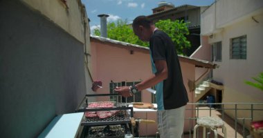 Afrika kökenli Amerikalı bir adam çatıdaki barbeküde biftek yapmak için çatal bıçak kullanıyor. Yemeği özenle hazırlıyor ve rahatça açık havaya odaklanıyor.