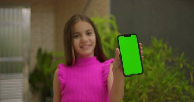 Genç kız kameraya yeşil ekran ve krom anahtar ile akıllı telefonu sunuyor, açık hava ortamında gülümsüyor, dijital ya da reklam amaçlı kişiselleştirilebilir görüntü sergiliyor