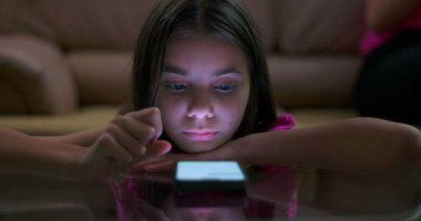 Parlak akıllı telefon ekranına odaklanırken kollarında dinlenen genç kız dijital bağlılık, teknoloji kullanımı ve modern çocukluk alışkanlıklarını gösteriyor