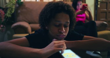 Oğlan cam masada akıllı telefona dalmış, kız koltukta telefona dalmış, duygusal izolasyonu, sosyal kopukluğu ve dijital bağımlılığı resmediyor.