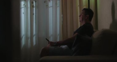 Kanepeye yaslanıp televizyon izleyen, uzaktan kumandayı elinde tutan, arka planda perdeler, rahat ev ortamları, rahat ev eğlencesi, samimi ve doğal an.