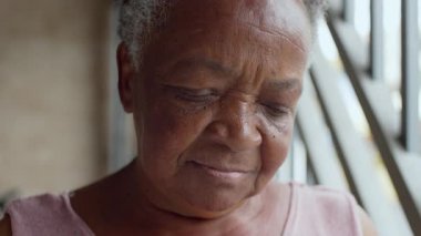 Yansıtıcı bir ifadeyle aşağı bakan yaşlı kadının yakın çekimi, gün ışığıyla aydınlanan yüzü, huzurlu bir tefekkür anında sokak garajında demir parmaklıkların yanında.