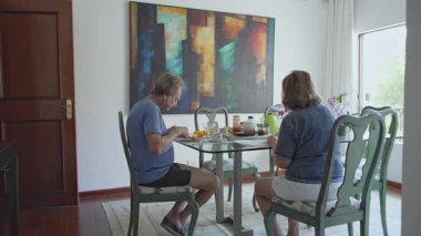 Evde kahvaltı yapan yaşlı bir çift, çeşitli yiyeceklerle birlikte cam bir masada oturuyorlar.