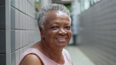 Yaşlı Afro Kadın, sessiz bir koridorda, duvarın önünde oturmuş sıcak bir şekilde gülümserken, Yüzündeki ifadeyle mutluluğu ve enerjiyi selamlıyor.