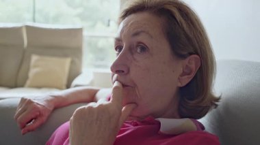 Koltukta oturan, elleri çenesinde ve yansıtıcı ifadesiyle yaşlı bir kadının yakın çekimi.