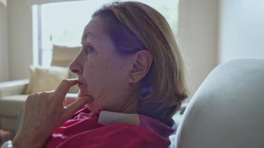 Düşünceli bir ifadeyle koltukta oturan yaşlı kadın, başka tarafa bakarken elini çenesine koyuyor.