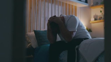 Orta yaşlı bir adam gece yatağında oturuyor, kafası ellere gömülmüş, duygu ve yorgunluktan bunalmış, kederi iletiyor, umutsuzluk, ve samimi ev ortamında sessiz bir umutsuzluk.