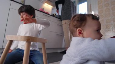 Büyük kardeş mutfakta bebeğin yanında oynarken, ebeveyn yemek pişirirken, sıcak ev ortamında günlük aile etkileşimlerini, birlikteliklerini ve evdeki bağlantılarını gösteriyor.