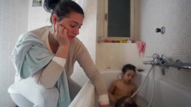 Anne küvetin üzerine eğilip iki çocuğu sıcak ve sıcak bir iç anda yıkıyor. Şefkat yorgunluğu ve yumuşak doğal ışıkta sevgi gösteriyor.