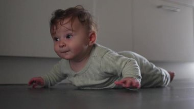 Bebek mutfak zemininde uzanmış başını kaldırıyor. İfade eden yüzüyle merak ve çaba gösteriyor. Yumuşak ev ışığı altında.