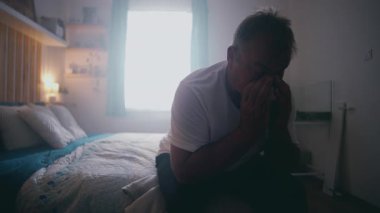 Yatakta oturan bir adam elleri ile yüzünü kapatıyor, bunalmış ve duygusal, derin üzüntü, stres ve umutsuzluk ifade ediyor samimi ev ortamında.