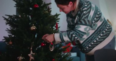 Anne sakin bir akşam ışığında Noel ağacına süsler ekliyor. Farkındalığı, basitliği ve şenlikli kış mevsimi için evi hazırlamanın sessiz neşesini ifade ediyor.