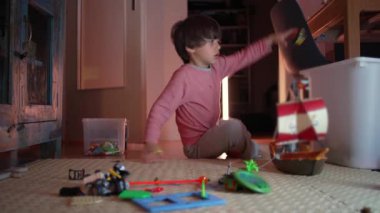 Genç bir çocuk yerde oturup oyuncakları topluyor, oyun saatinden sonra onları plastik bir kaba koyuyor.