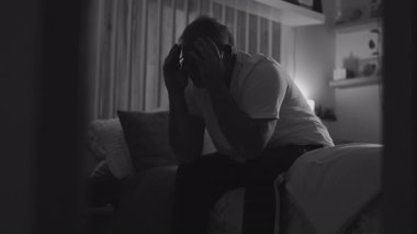 Orta yaşlı bir adam yatağında oturmuş gözlüklerini çıkarıyor ve yüzünü yorgunluk ve umutsuzlukla örtüyor. Derin pişmanlık ve duygusal karmaşa içinde loş bir odada.