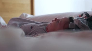 Bebek yatakta uzanıyor, kumaş kayışını inceliyor ve gün ışığında inceliyor. Merak ve eğlence dolu konsantrasyonunu nazik, samimi bir iç atmosferde gösteriyor.