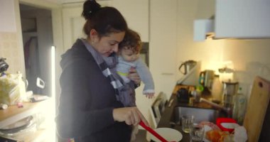 Anne bebek tutarken yemek pişirir, ev işlerini ve çocuk bakımını üstlenir, sakin bir şekilde odaklanır, sabırlı olur ve evde günlük anneliğin duygusal gücünü ifade eder..