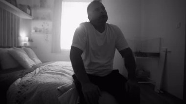 Yatakta tek başına oturan bir adam, siyah ve beyazı yansıtırken yorgunluğunu, umutsuzluğunu ve duygusal mücadelesini ifade ederek umutsuzluğa kapılmış bir şekilde yukarıya bakıyor.