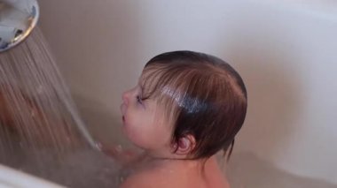 Bebek duşun altında oturuyor, sakin bir ifade evde sıcak banyo zamanında merak ve rahatlık gösteriyor.