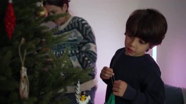 Noel ağacına asılan çocuk süsü, yanında annesi olan, samimi aile anı neşe, konsantrasyon ve şenlikli ev atmosferinin sıcaklığını gösteren.