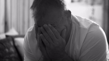 Orta yaşlı bir adam, elleri ve elleri ile yatakta oturuyor, siyah ve beyaza yakalanıyor. Saf üzüntü, yalnızlık ve duygusal yorgunluğu samimi ve gerçekçi bir ortamda ifade ediyor.