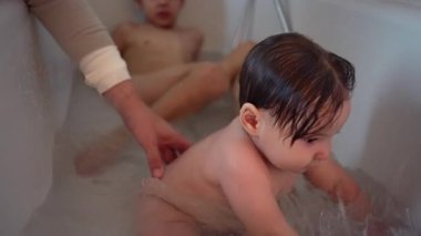 Bebek küvette oynuyor, büyük kardeş yakınlarda, hassas aile banyosu zamanı sıcak evde bakım, masumiyet ve erken çocukluk sevincini gösteriyor.