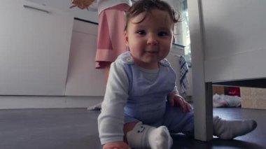 Bebek mutfak zemininde kameraya yakın duruyor, merakla ve dikkatle ileri bakıyor, merceğe uzanıyor ya da dokunuyor gibi, erken duyusal bağlılık ve masumiyeti gösteriyor.