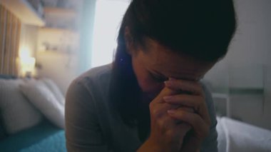 Başı eğik ve elleri sımsıkı bağlı bir şekilde yatakta tek başına oturan kadın, gün ışığında duygusal yorgunluğunu, yansımasını ve sessiz kederini ifade ediyor.