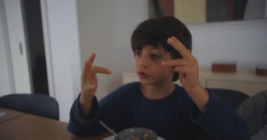 Küçük çocuk masada oturup parmaklarıyla sayarken kardeşi izliyor ve gülümsüyor. Merakını, öğrendiklerini ve paylaşılan aile anlarının mutluluğunu ifade ediyor.