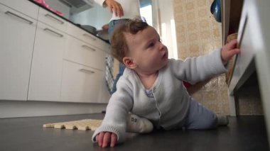 Ebeveyn yakınlarda yemek pişirirken, mutfak zemininde emekleyen bebek aile ortamında merak ve erken keşif gösteriyor.