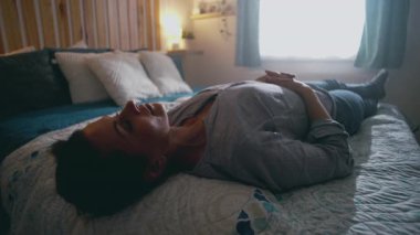 Kadın gün ışığında yatakta sessizce uzanıyor, elleri göğsünde dinleniyor, yorgunluk, yalnızlık ve huzur dolu bir ortamda duygusal duraksama yansıtıyor.