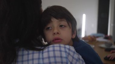 Genç bir çocuk mutfakta annesinin omzunda dinleniyor, gözleri yarı açık, rahatlık, yorgunluk ve aile rutini sırasında sessiz duygusal bağ gösteriyor.