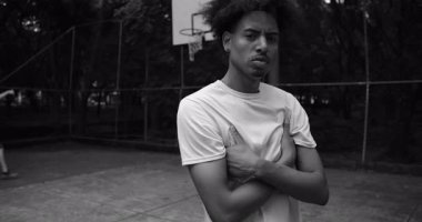 Açık hava basketbol sahasında kendine güvenen Afro-Amerikalı bir adam kollarını kavuşturmuş, ciddi bir ifade takınıyor, sokak oyunundan sonra sakin, tek renkli bir portre.