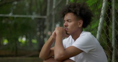 Genç Afrikalı bir adamın basketbol sahasında çitin kenarında oturması, odaklanma, yansıma ve iç kararlılığını gösteren düşünceli bir ifade.