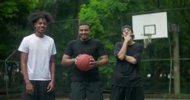 Üç genç adam gülüyor ve dışarıda basketbol sahasında rahatlıyor, ağaçlarla çevrili gündelik kentsel spor ortamında arkadaşlıklarını, neşelerini ve arkadaşlıklarını ifade ediyorlar.