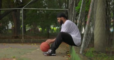 Genç Afrikalı kökenli bir adam çitin yakınındaki açık basketbol sahasında oturuyor. Teneffüste düşünceli bir şekilde basketbol oynuyor. Odaklanma, yansıma ve alıştırma anı.