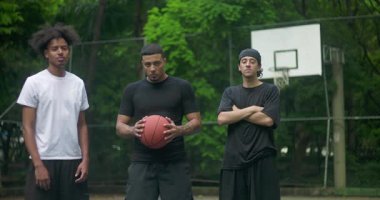 Arka planda potası olan, takım çalışmasını, odaklanmayı ve şehir sporları kültüründe disiplini temsil eden, ciddi ve kendinden emin üç genç adam dışarıda basketbol sahasında duruyor.