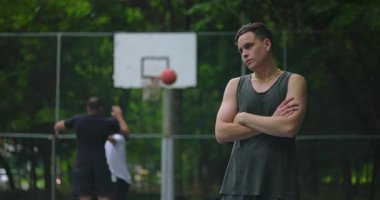 Latin asıllı genç bir adam basketbol sahasında kollarını kavuşturmuş, kuşkucu bir ifadeyle yan bakıyor, kendine güvenini ve kurnazca meydan okuyor.