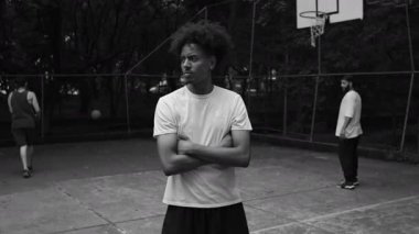 Açık havada tek renkli basketbol sahasında duran, kollarını kavuşturan, kentsel spor ortamında yoğunluk ve kendine güveni olan Afro-Amerikalı bir adam.
