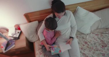 Anne oğlunu kucağına alıp uyku masalı okurken şefkatli bir aile anı, eğitim ve ev ortamında duygusal bir bağ gösteriyor.