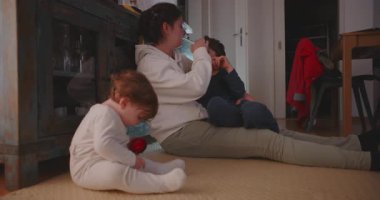 Anne iki çocukla yerde oturuyor, yanında bebek oynarken büyük oğlunu teselli ediyor, evde sıcaklığı, ilgiyi ve aile bağlarını yakalıyor.